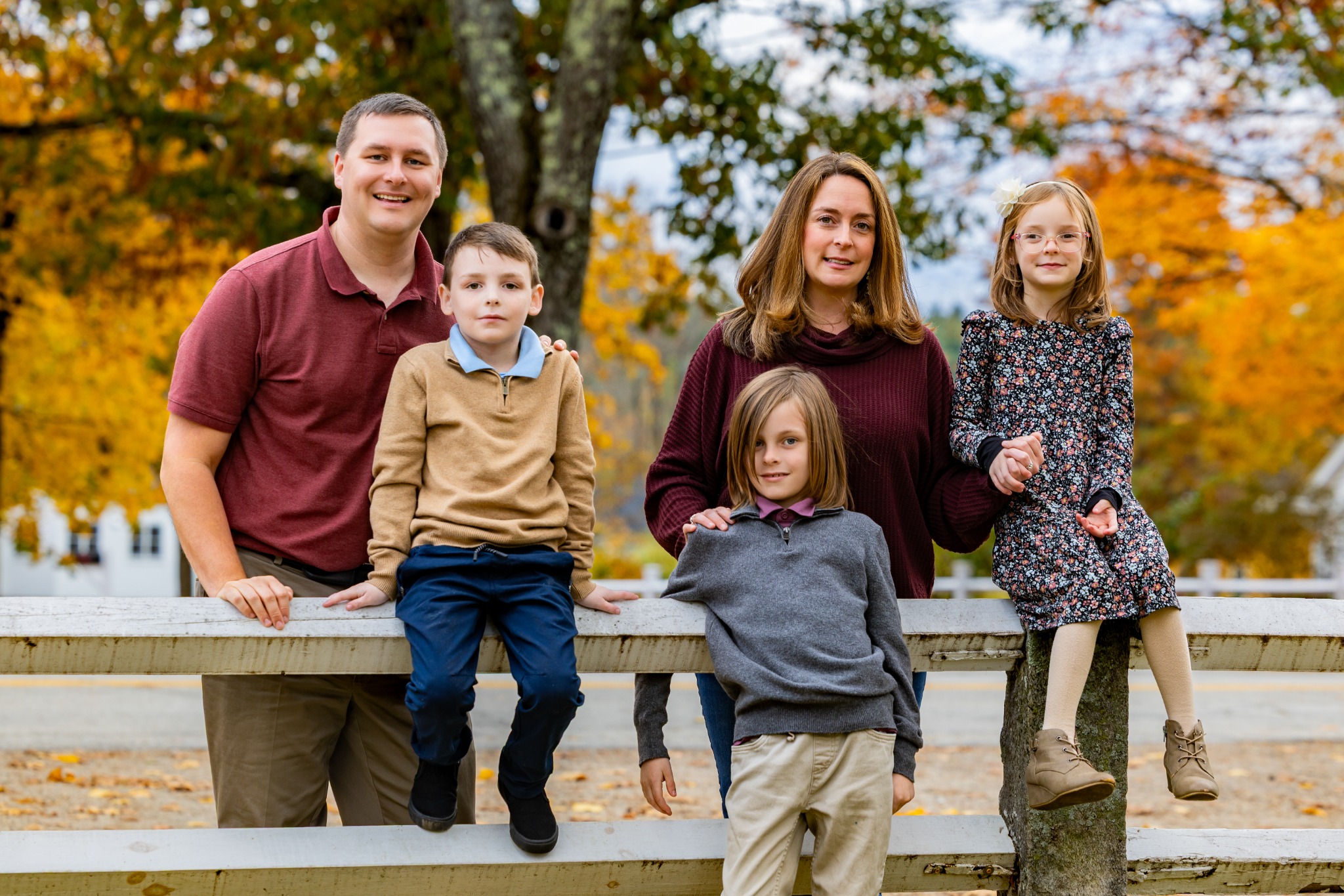 A family of five together beside a white fence surrounded by autumn foliage